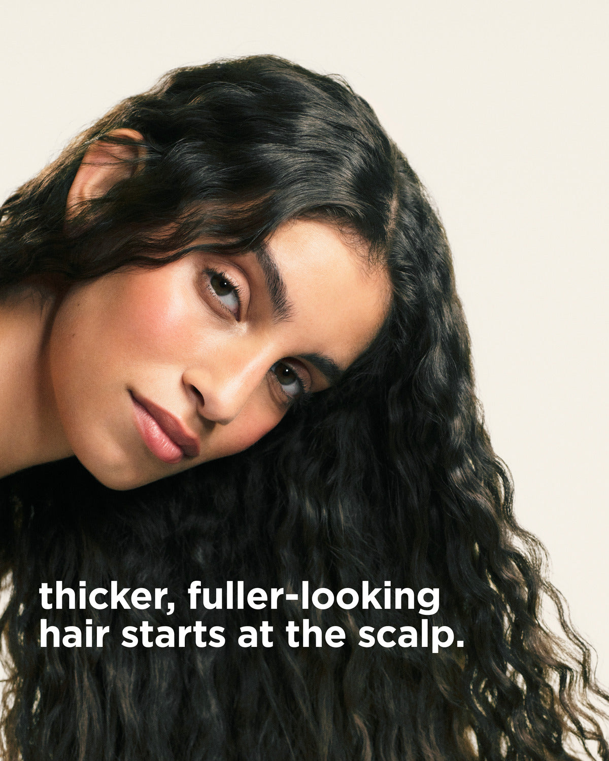 Close-up of a woman with long, dark, wavy hair looking toward the camera, with the text 'thicker, fuller-looking hair starts at the scalp.' overlaid at the bottom left on a light beige background.