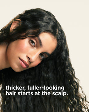 Close-up of a woman with long, dark, wavy hair looking toward the camera, with the text 'thicker, fuller-looking hair starts at the scalp.' overlaid at the bottom left on a light beige background.